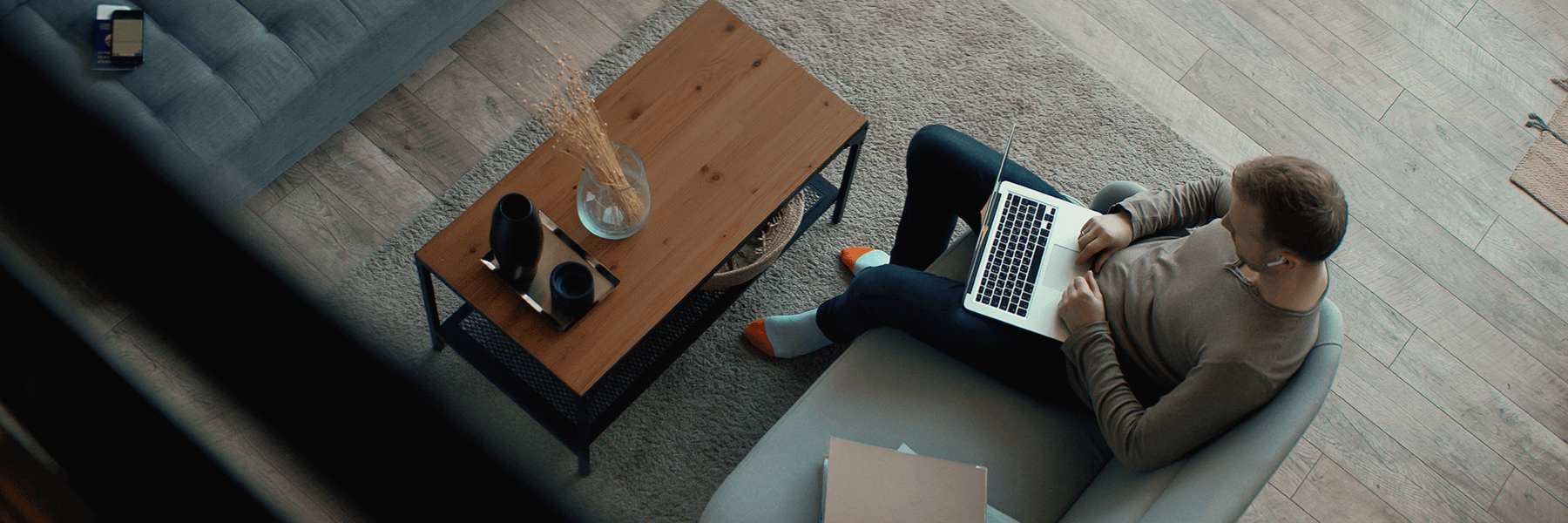 Man in sweats and socks sits at his couch doing work on his laptop computer