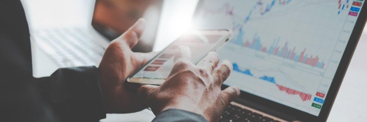 person on laptop and phone, screens showing financial data